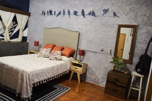 Dormitorio acogedor en LLUNA Oaxaca con cabecero beige, almohadas naranjas, mesita de noche de madera y arte mural de pájaros en alambre