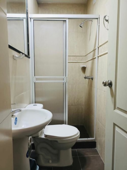 Pequeño baño del hotel Casa Mango en Cali con inodoro, lavamanos y ducha con puertas de vidrio marco blanco