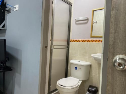 Baño del hotel Casa Mango en Cali con inodoro, lavamanos, espejo y cabina de ducha con borde decorativo de azulejos