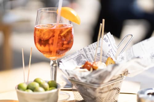 Aperitif cocktail with orange slice at YOUnique Boutique Rooms Milano, featuring olives and newspaper-wrapped snacks