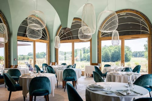 Le Prieuré Du Preux dining room with arched windows, pendant lights, teal chairs, and countryside views in Sépeaux, France.
