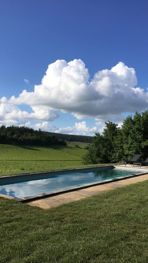 Piscine rectangulaire dominant les vignobles verdoyants et les collines boisées sous un ciel bleu parsemé de nuages blancs aux Maisons de Chamirey