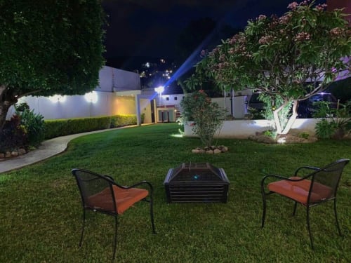 Vista nocturna del jardín de Casa Palmeras con fogata, asientos, césped exuberante y arquitectura blanca moderna