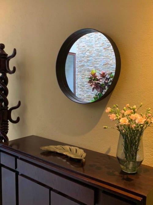Cómoda de madera oscura con espejo negro redondo, pluma dorada y claveles naranjas en jarrón de cristal contra pared amarillo mostaza