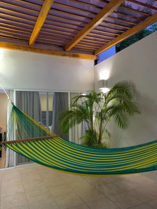 Hamaca de rayas verdes y amarillas en el patio de azulejos de la cabaña Casa Palmeras con palmera en maceta y pérgola de madera