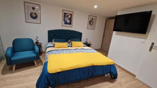Chambre moderne d'appartement avec tête de lit bleue, literie jaune, fauteuil turquoise, décoration murale et TV écran plat