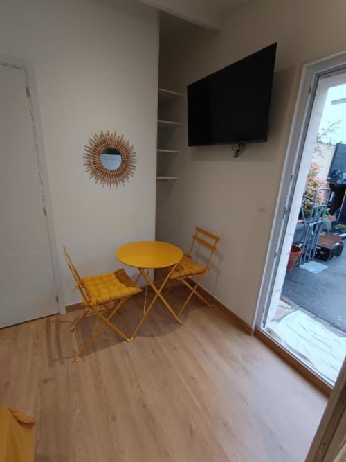 Séjour lumineux avec table ronde jaune, deux chaises, miroir soleil, téléviseur mural et porte vitrée donnant sur balcon