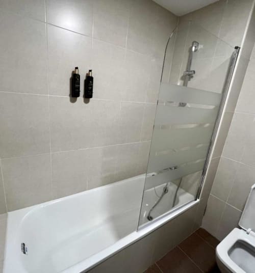 Baño moderno con azulejos blancos, bañera, cabina de ducha de cristal y servicios en el apartahotel AlmaMaterSalamanca.