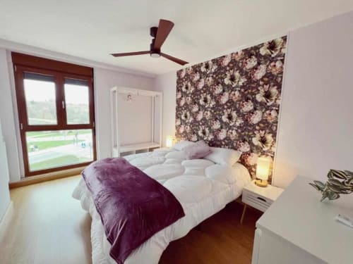 Dormitorio moderno en AlmaMaterSalamanca con pared de flores, ropa de cama blanca, manta púrpura y vista a la ciudad