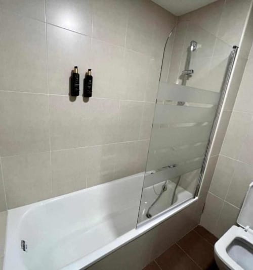 Baño moderno con azulejos blancos, ducha de cristal sobre bañera y piso de azulejos oscuros en Apartamentos