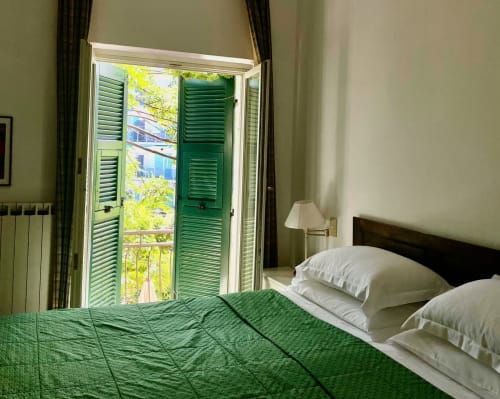 Chambre lumineuse à l'Hôtel Beau Rivage Alassio avec portes-fenêtres aux volets verts, oreillers blancs et vue sur le jardin luxuriant