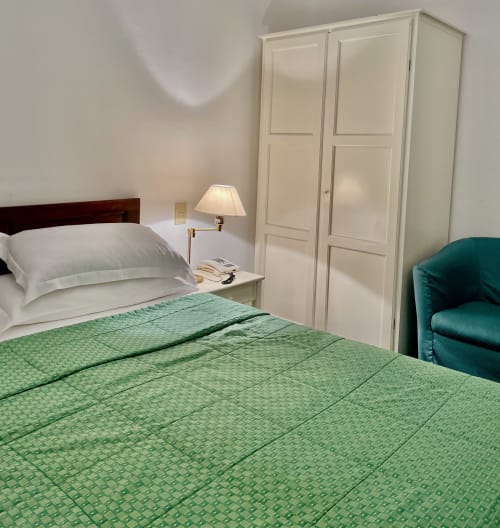 Klassisches Zimmer im Hotel Beau Rivage Alassio mit grüner Bettwäsche, weißem Kleiderschrank, türkisfarbenem Sessel und Nachttischlampe