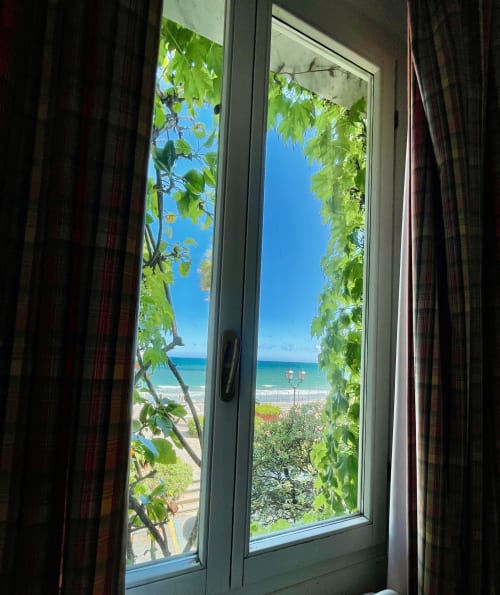 Blick aus dem Zimmer des Hotel Beau Rivage mit Meerblick, Strand, türkisblauem Wasser und üppigen grünen Ranken, die das Fenster rahmen, Alassio