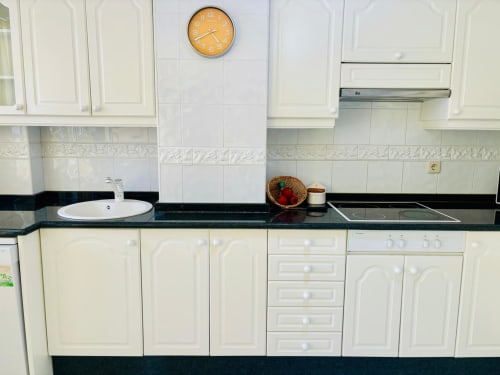 Cocina moderna con armarios color crema, encimera negra, azulejos blancos, reloj mural amarillo y electrodomésticos en Baiona