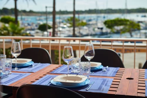 Mesa de comedor frente al mar en Colonia de Sant Jordi con vistas al puerto, cubiertas azules, copas de vino y barcos