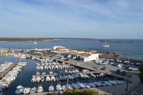 Port esportiu amb embarcacions blanques amarrades a la pasejada marítima de Colonia de Sant Jordi, Espanya, mar blau cristal·lí i edificis costaners