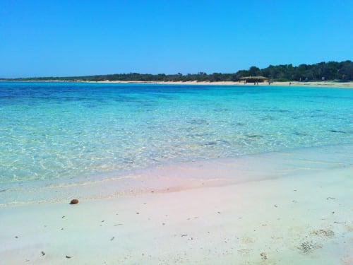 Aigües turqueses pristines i platja d'arena blanca a Colònia de Sant Jordi amb cel blau clar i vegetació litoral distant