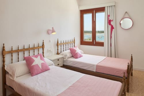 Dormitori lluminós amb dos llits de fusta, coixins amb estrelles rosades i vistes al mar a través de finestres amb marc vermell a Colonia de Sant Jordi