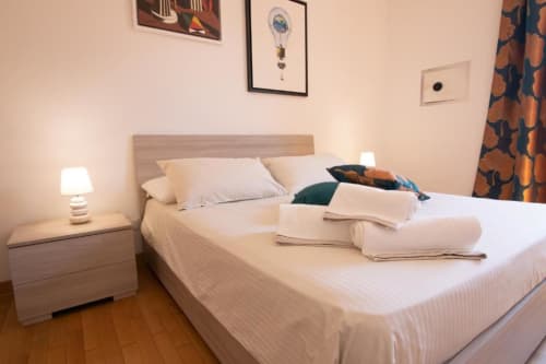 Camera luminosa a Dimore Verona con letto bianco, comodino in legno, lampada e vista sulla città dalla finestra