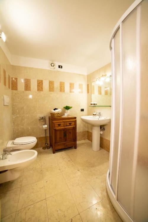 Bagno spazioso a Dimore Verona con piastrelle beige, lavabo su piedistallo, wc, mobile in legno e box doccia