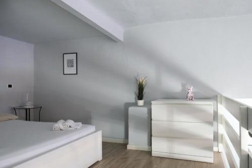 Camera da letto in soffitta minimalista con arredi bianchi, pianta in vaso e luce naturale dalla finestra a lucernario.