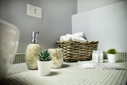Vanità moderna del bagno a Dimore Verona con dispenser di sapone decorativo in oro, pianta succulenta, cesto intrecciato e asciugamani bianchi