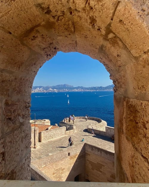 Vue à travers une ancienne arche de pierre du port de Marseille avec voiliers et paysage urbain côtier au-delà