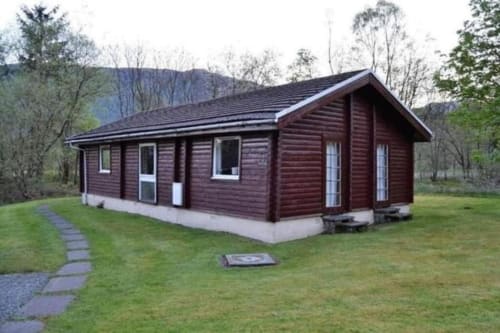 Modern dark red log cabin at Grouse Lodge in Dunoon with stone foundation, surrounded by green lawn and trees