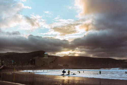 Surfers enjoy Atlantic waves at Arguineguín beach with dramatic golden-hour sky and beachfront hotel building