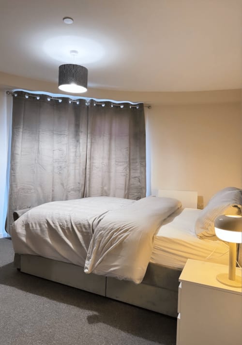 Modern bedroom at Cila Property Manchester hotel with bed, taupe curtains, ceiling light, and side table lamp