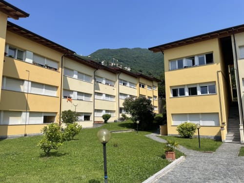 Yellow apartment buildings at Varenna by Foot in Italy with green courtyard, mountains, and blue sky
