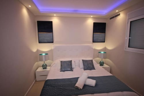 Modern bedroom in Marbella penthouse with blue ambient ceiling lighting, white headboard, dark accent pillows, and