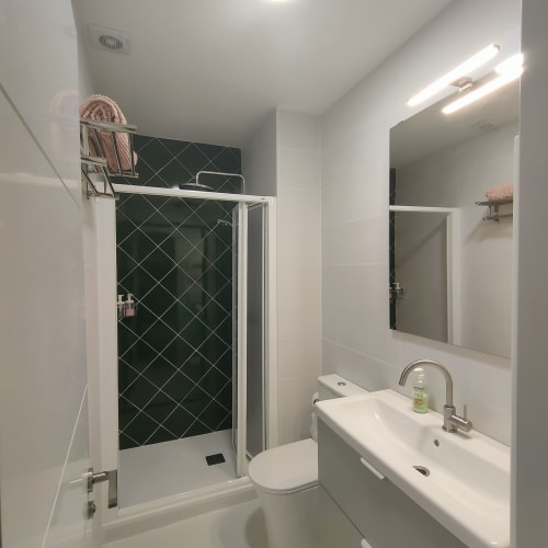 Baño moderno del apartamento Megaleon en Segovia con ducha de azulejos negros en forma de diamante, vanidad blanca e iluminación del espejo.