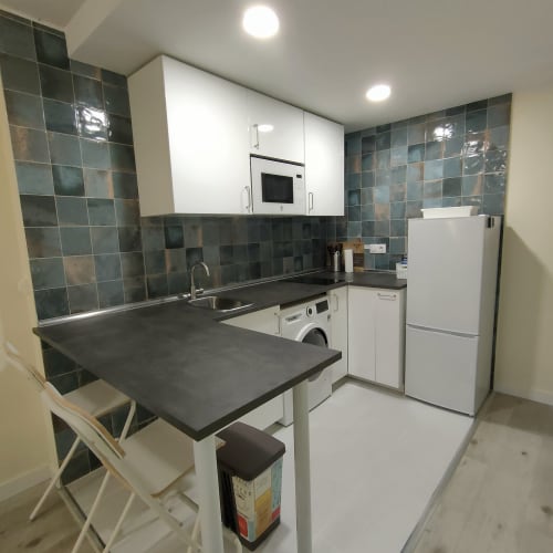 Cocina moderna del hotel Megaleon en Segovia con azulejos oscuros, muebles blancos, encimera negra y mesa de comedor.