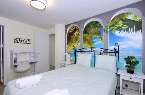 Dormitorio luminoso del hotel Megaleon en Segovia con mural tropical, cama de hierro forjado y papel tapiz con vista al océano.