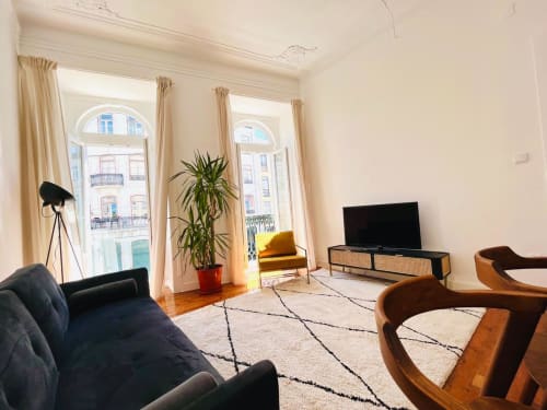 Sala de estar moderna no Royal Prince Garden Apartments em Lisboa com sofá preto, cadeiras de madeira, TV, plantas e janelas em arco