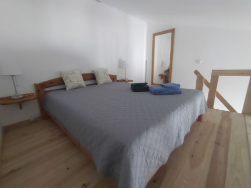 Quarto espaçoso na Casa do Pão com cama em madeira, roupa de cama cinza, espelho e piso em madeira clara