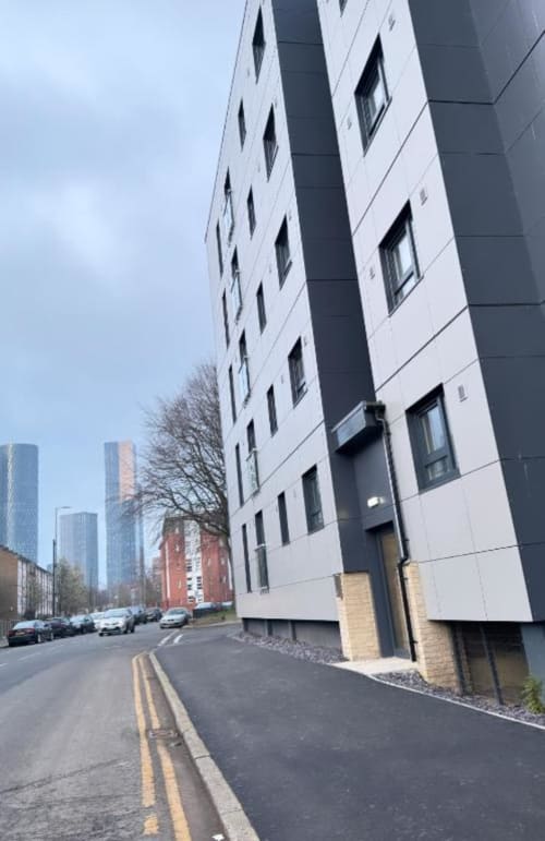 Modern white apartment building in Manchester with dark vertical panels, street view with parked cars and city skyline