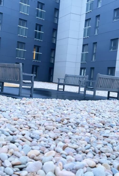 Pebble beach patio with modern blue apartment building at Cila Property Manchester, featuring wooden deck and garden view