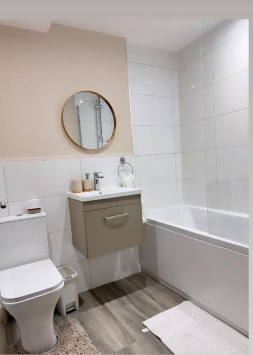 Modern bathroom at Cila Property Manchester with white tiles, beige walls, round mirror, olive vanity, and bathtub with glass