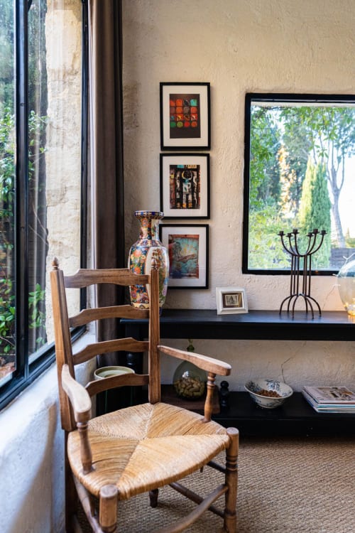 Coin de lecture cosy au Mas F à Saint Rémy de Provence avec fauteuil en bois, tableaux encadrés et vue sur le jardin