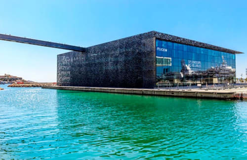 Bâtiment muséal contemporain sur le front de mer de Marseille avec les eaux turquoise du port et un ciel dégagé