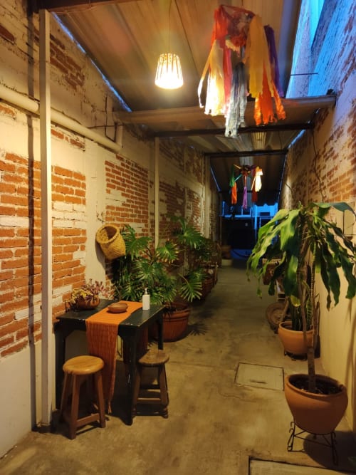 Patio del apartamento LLUNA en Oaxaca con muros de ladrillo expuesto, plantas en macetas, mesa de comedor, telas colgantes e iluminación cálida