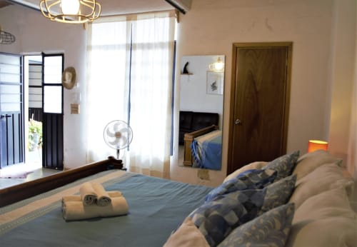 Dormitorio luminoso en apartamento LLUNA de Oaxaca con ropa de cama azul, puerta de madera, ventilador y luz natural de las ventanas