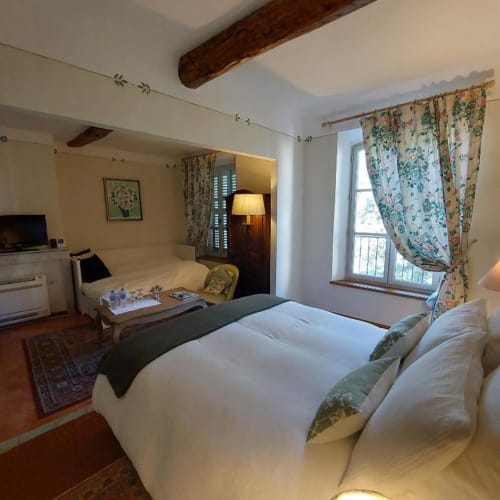 Chambre lumineuse à La Cordeline avec lit queen, rideaux fleuris, poutres en bois et vue sur la ville