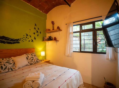 Dormitorio luminoso en tonos amarillo y naranja del apartamento LLUNA en Oaxaca, con cama de madera, arte de plumas en la pared, estantes y gran ventana