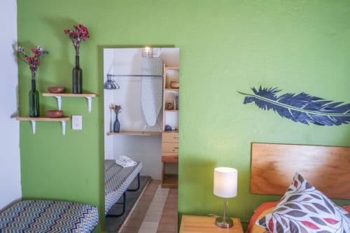 Dormitorio verde brillante en el apartamento LLUNA de Oaxaca con cama de madera, repisas flotantes, flores rosas y arte mural de plumas azules