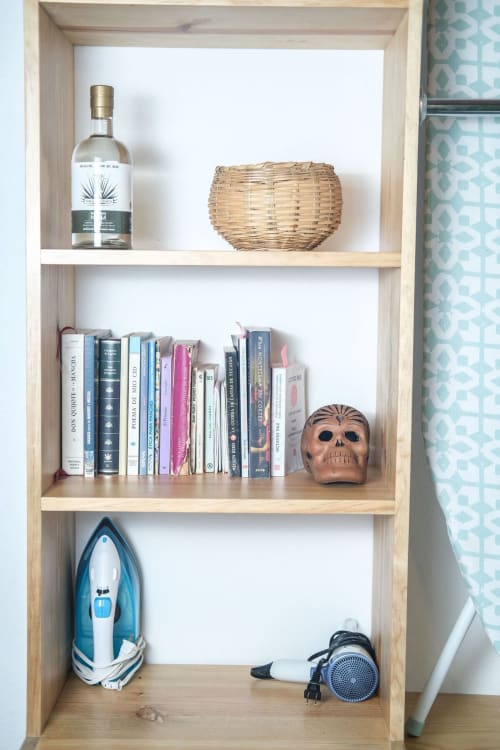 Estante de madera clara en apartamento LLUNA con libros, calavera decorativa, cesta tejida, botella de licor, plancha y secador de pelo