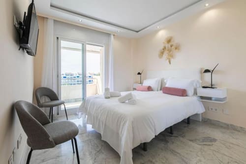 Modern bedroom at Marbella Home Rentals Marina Banús with city views, white bedding, gray chairs, balcony access, and