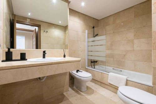 Modern bathroom with beige marble tiles, double vanity, toilet, and shower-tub combo at Marina Banús apartment in Marbella